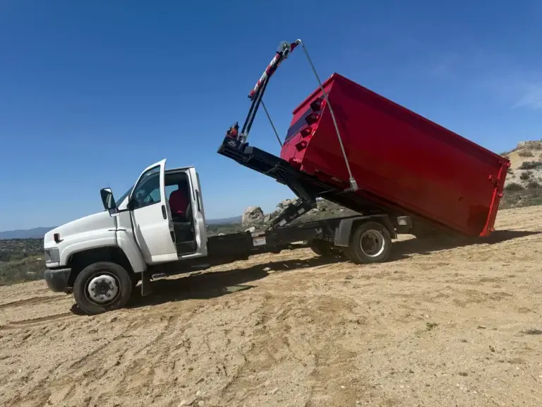 A white pickup truck with its driver’s door open is attached to a red dump truck. The dump bed is raised by a hydraulic crane, lifting part of the red container. The scene is on a dusty, sloped dirt area with hills in the background under a clear blue sky.