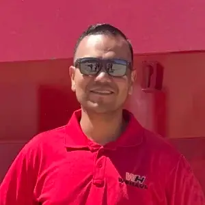 A man wearing a red polo shirt and dark sunglasses smiles at the camera. He has short hair and is standing outdoors in front of a red background.
