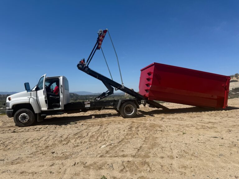 A white flatbed tow truck on a dirt hill, with a black crane arm lifting something, and a large red dumpster tilted at the back. Clear blue sky in the background.