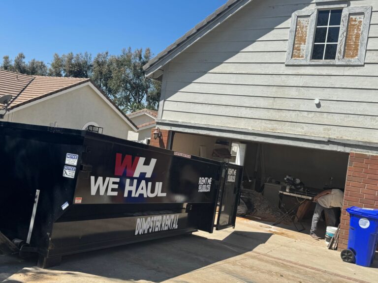 A black dumpster with “WE HAUL” branding and “DUMPSTER RENTAL” text sits open on a residential property beside a driveway and garage, with trash bags and bins nearby.