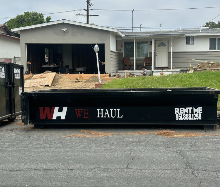 A black dumpster/trailer parked in a residential driveway with bold red and white text reading “WH WE HAUL” and “RENT ME” followed by the phone number “951-888-1258.”