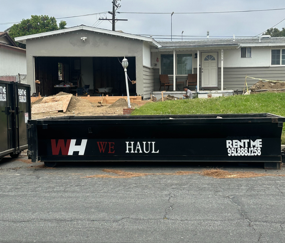 A black dumpster/trailer parked in a residential driveway with bold red and white text reading “WH WE HAUL” and “RENT ME” followed by the phone number “951-888-1258.”
