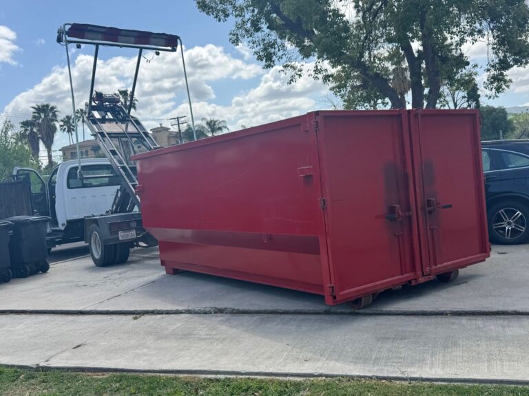 A red open-top dump truck with a hydraulic lift mechanism is parked at a curb in a sunny outdoor area with trees and other vehicles nearby.