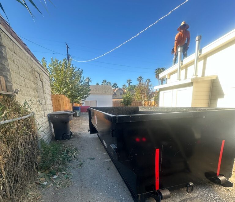 Backyard scene with a worker standing near a large black dumpster-style container, trees and fencing along the sides, and a clear blue sky.