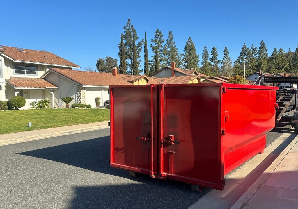 A bright red dumpster with double front doors sits on a black trailer along a residential street, with houses, tall trees, and a clear blue sky in the background.
