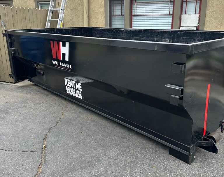 A black roll-off dumpster with "WH WE HAUL" logo parked on asphalt, with a wooden fence and building in the background.