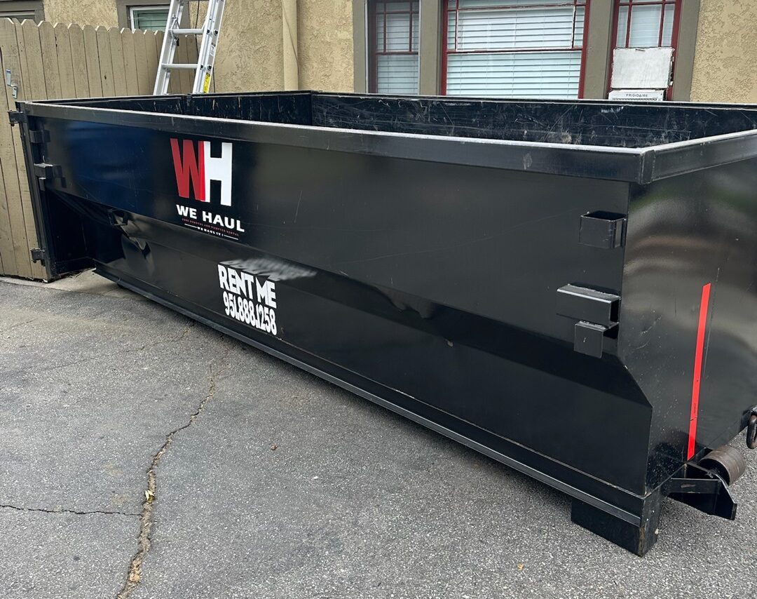A black roll-off dumpster with "WH WE HAUL" logo parked on asphalt, with a wooden fence and building in the background.