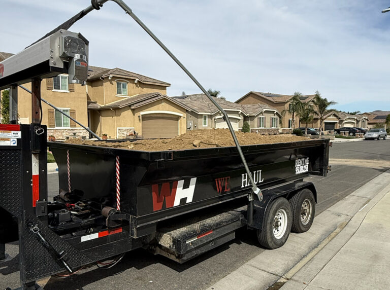A black dump trailer filled with dirt and debris sits parked on a residential street. The trailer features white tires and a metal arm extending over the load. Its side displays "WE HAUL" in red and white lettering along with "RENT ME" in white text. In the background, light brown stucco houses with tile roofs and solar panels line the street, with palm trees and a partly cloudy sky overhead.