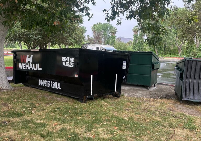 A large black dumpster with white lettering sits on grass beneath a tree. The dumpster reads "WH WEHAUL" on the left, "RENT ME 951.888.1258" on the upper right, and "DUMPSTER RENTAL" along the bottom. In the background on wet pavement, two green dumpsters are visible, along with lush trees and a white vehicle or trailer.