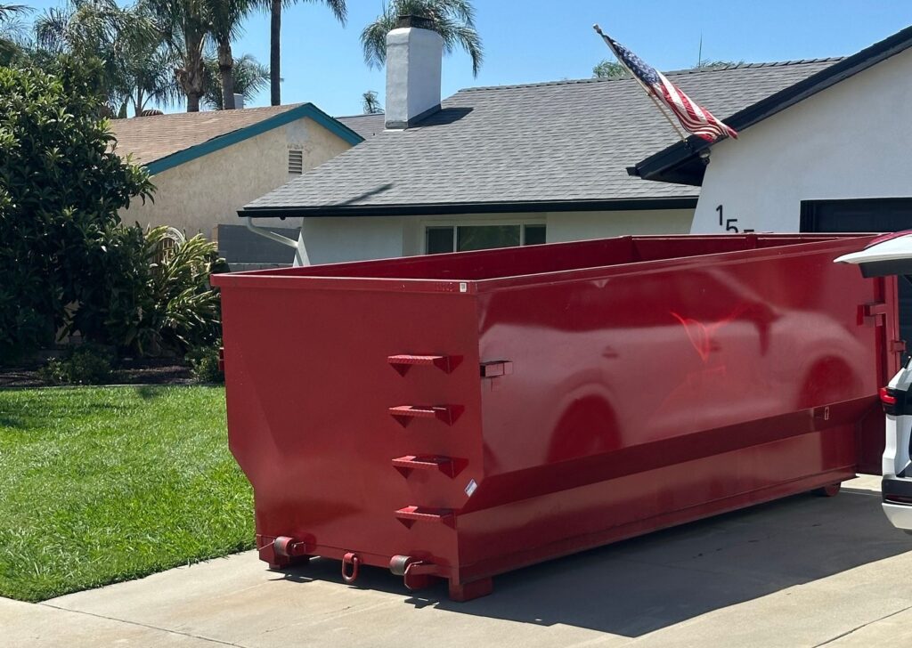 A large red dumpster parked on a concrete driveway in front of a white single-story house with a gray roof, white chimney, and an American flag hanging from the side. A smaller tan house with blue trim is visible in the background, with green grass and trees under a blue sky.