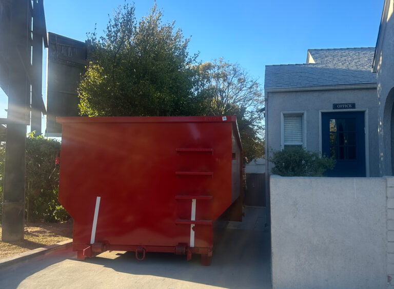 A large red dumpster sits on a cracked concrete surface under a clear blue sky, with a small building featuring a blue door and an "OFFICE" sign behind it, and trees with a wooden fence or structure to the left.