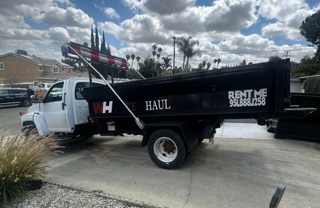 A white work truck with a black dump bed is parked on a residential concrete driveway. The truck's side reads "WH VE HAUL" along with "RENT ME" and the phone number 951.888.1258. Red and white striped safety markers are mounted on top of the truck bed. The background features houses, palm trees, and an overcast sky.