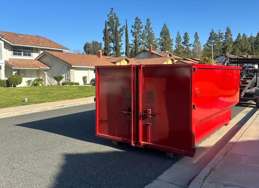 The image shows a red dumpster with open doors parked on the right side of a residential road, with a flatbed truck behind it and houses with green lawns across the street under a blue sky.