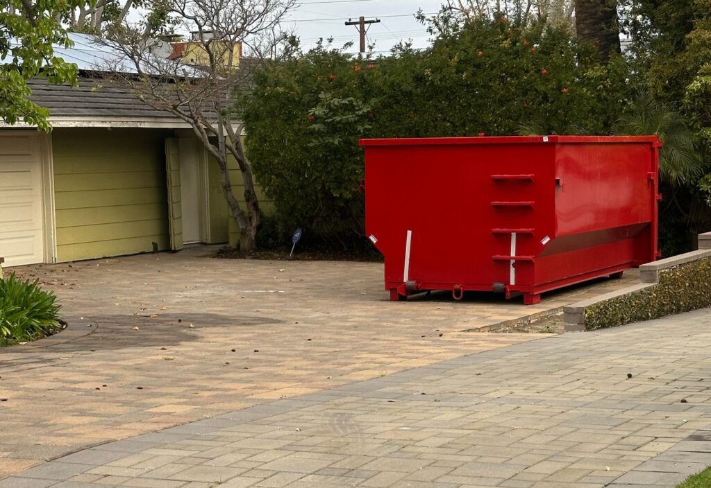 A bright red dumpster sits on a patterned stone driveway next to a light green garage with white doors. Behind the dumpster is a dense green hedge and trees, with power lines visible against the sky.