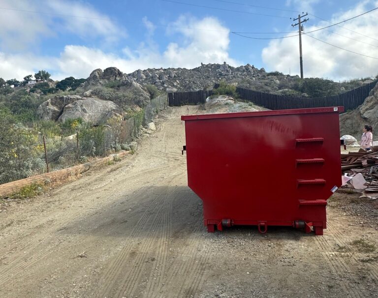 A large red dumpster sits on a dirt road in the foreground, with a person standing to its right near a pile of scrap metal. Behind it, a hill rises with scattered rocks and boulders amid dry vegetation. A wooden fence runs along the slope, a utility pole stands to the right, and a house is visible near the hilltop under a blue sky with white clouds.