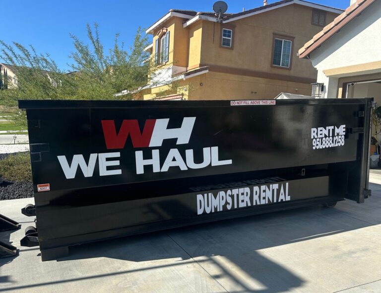 A black dumpster labeled "WH WE HAUL" with "DUMPSTER RENTAL" and the phone number 951.888.1258 sits on a driveway in front of a two-story tan stucco house. A warning reads "DO NOT FILL ABOVE THIS LINE." Blue sky and green shrubs are visible in the background.