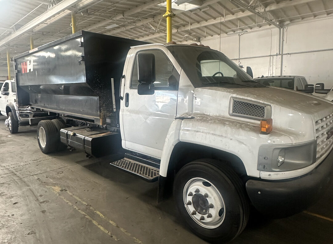 A dirty white dump truck with a black rectangular dump bed parked inside a warehouse or garage, with a concrete floor and high ceiling supported by metal beams. Another similar white truck with a black dump bed is partially visible behind it.