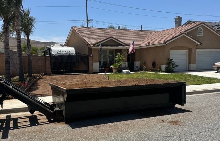 A dark gray roll-off dumpster filled with dirt sits on a paved street, partially lowered from a truck with its lifting arm visible in the foreground. In the background, a light brown stucco house with a tiled roof and green lawn is visible, with an American flag hanging from the porch. A white RV marked "VORTEX" is parked behind a dark metal gate to the left. Several palm trees line the property, and power lines cross a bright blue sky.