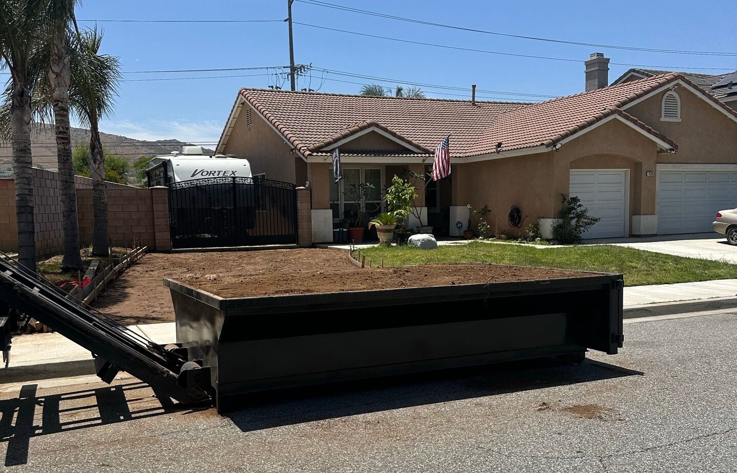 A dark gray roll-off dumpster filled with dirt sits on a paved street, partially lowered from a truck with its lifting arm visible in the foreground. In the background, a light brown stucco house with a tiled roof and green lawn is visible, with an American flag hanging from the porch. A white RV marked "VORTEX" is parked behind a dark metal gate to the left. Several palm trees line the property, and power lines cross a bright blue sky.