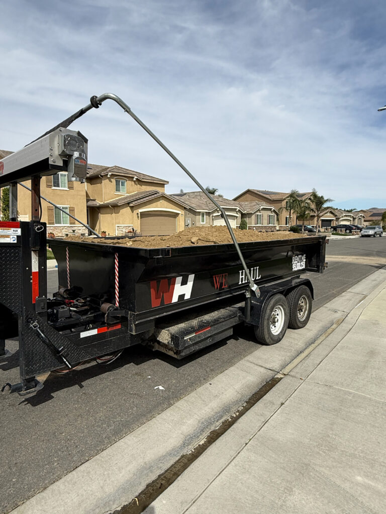 A black dump trailer filled with dirt, parked on a residential street. The trailer features a white "WH" logo with "WE HAUL" in red and white lettering on its side, and a "RENT ME" sticker on the back. A metal lifting arm extends above the trailer. In the background, suburban houses with light stucco and brown roofs line the street, with palm trees visible in the distance.