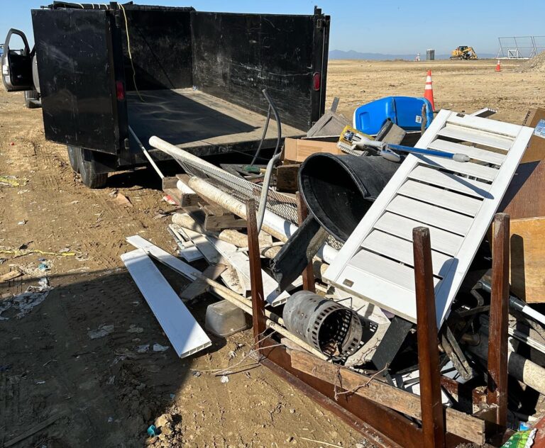 Construction debris and trash scattered on a dusty dirt lot in the foreground, including a pair of white louvered shutters, a black flexible hose, wooden planks, and a perforated metal cylinder. A black dump truck sits in the middle ground with a construction excavator and orange traffic cones visible further back under a clear blue sky.