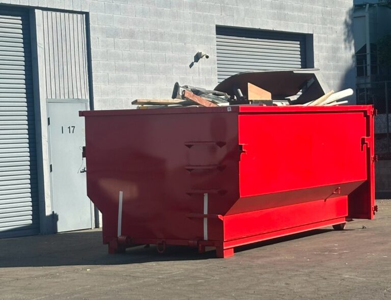 A bright red roll-off dumpster filled with construction debris—wooden planks, cardboard, and white pipes—sits on asphalt outside a light gray cinder block building. A gray garage door with horizontal slats and a door marked "1 17" are visible beside it. Sunlit outdoor setting.