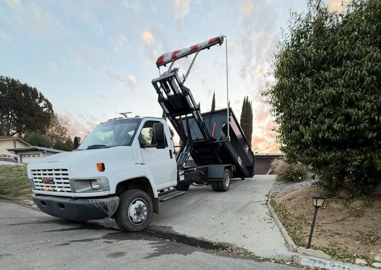 A white GMC dump truck with a raised hydraulic skip bin is parked on a residential driveway at an angle. Soft golden light, dirt, fallen leaves, and a small garden lamp complete the scene.