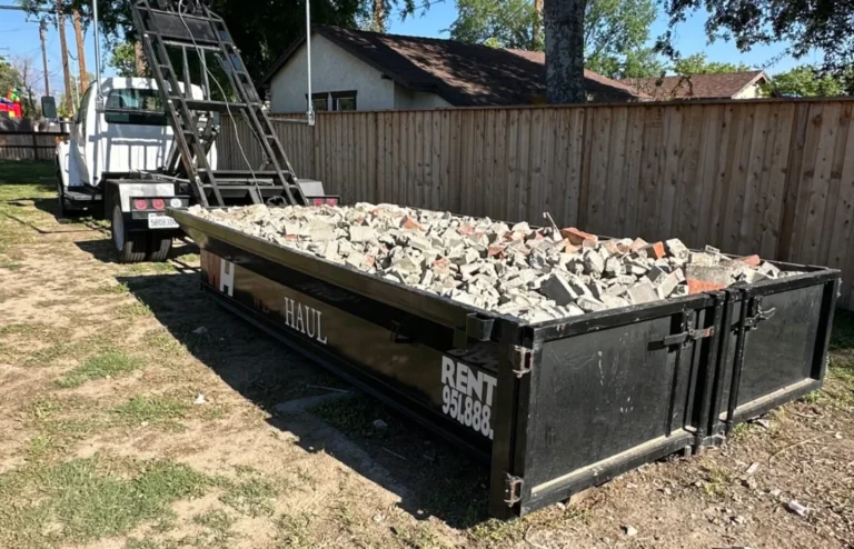 White roll-off truck backed up to a black dumpster filled with broken concrete and bricks in a grassy backyard near houses.