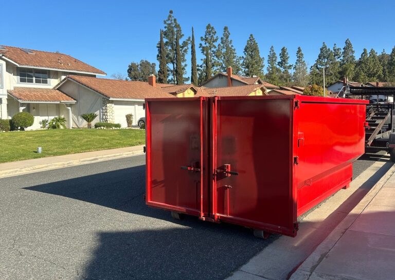 A red dumpster sits on a residential street under a sunny sky, with houses with orange-tiled roofs and green trees in the background.