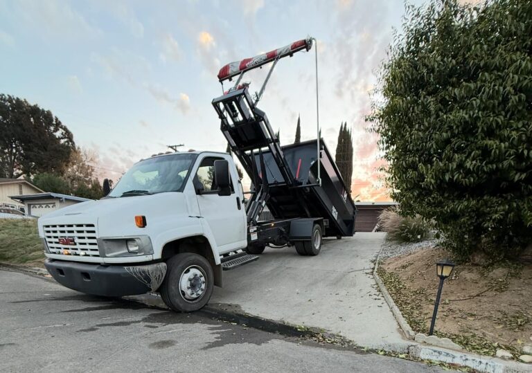 A white GMC dump truck with its bed raised carries a large black dumpster partially extending onto a driveway. A house, trees, bushes, and a sunset-tinged cloudy sky fill the background. A small black lamp post stands to the right.