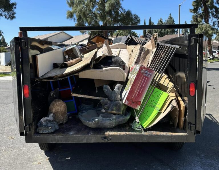 A rear view of a trailer piled high with assorted discarded items, including wooden planks, an old piece of furniture, cardboard boxes, and oversized gray stone blocks, set outdoors under a clear blue sky with residential houses and trees in the background.