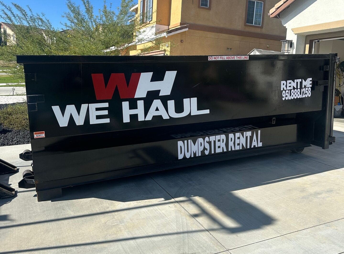 Black dumpster with "WH WE HAUL" logo and "DUMPSTER RENTAL" on its side, parked on a sunny driveway in front of a house. Red label reads "DO NOT FILL ABOVE THIS LINE." Contact number: 951.888.1258.