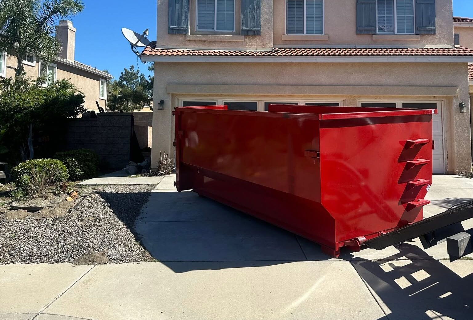 A bright red dumpster sits on the concrete driveway of a two-story beige house. The house has a garage door, two shuttered windows, and a satellite dish on the roof.