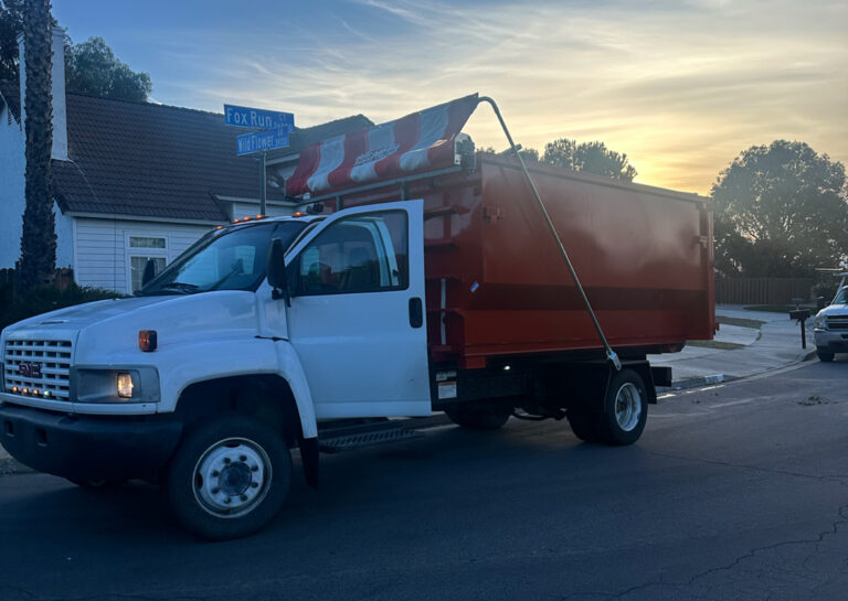 White dump truck with orange open-top container parked on a street at dusk, driver's door ajar, blue "Fox Run Wildflower" sign, house, trees, colorful sky