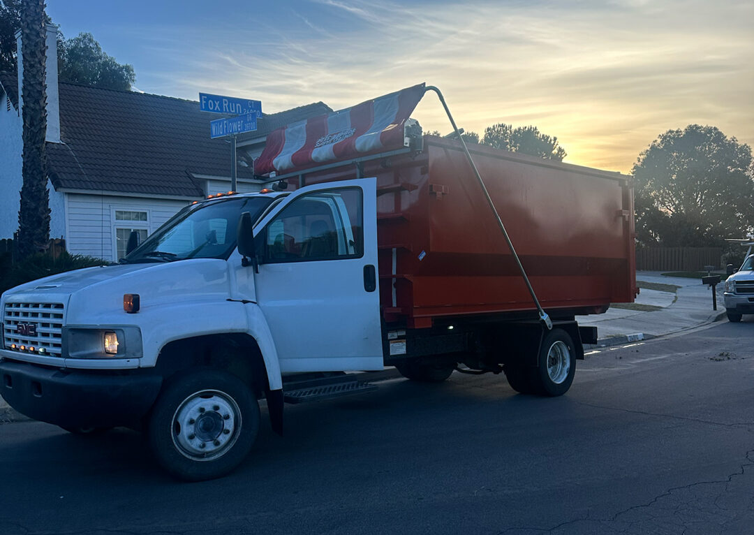 White dump truck with orange open-top container parked on a street at dusk, driver's door ajar, blue "Fox Run Wildflower" sign, house, trees, colorful sky