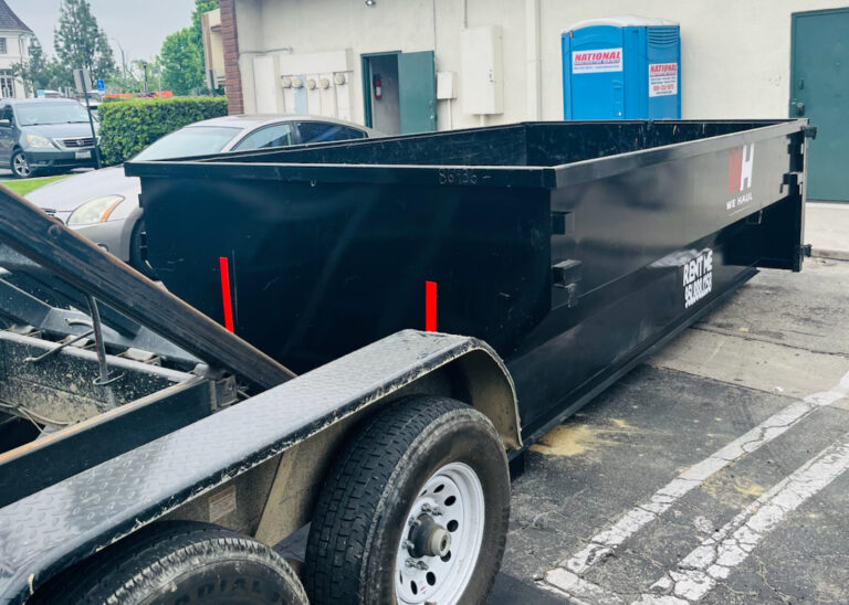 "A large black dumpster with red reflective strips sits on an asphalt lot, attached to a wheeled trailer for transport. In the background, a light-colored building with a red tiled roof stands behind a blue portable toilet labeled 'NATIONAL.' Several cars are parked in the distance, with green trees and bushes surrounding the area.