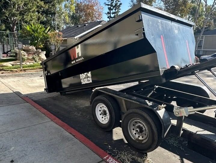 A large black dumpster labeled "WE HAUL" and "RENT ME" with a phone number, tilted on a flatbed trailer being loaded or unloaded, with green trees and residential buildings in the background under a clear sky.