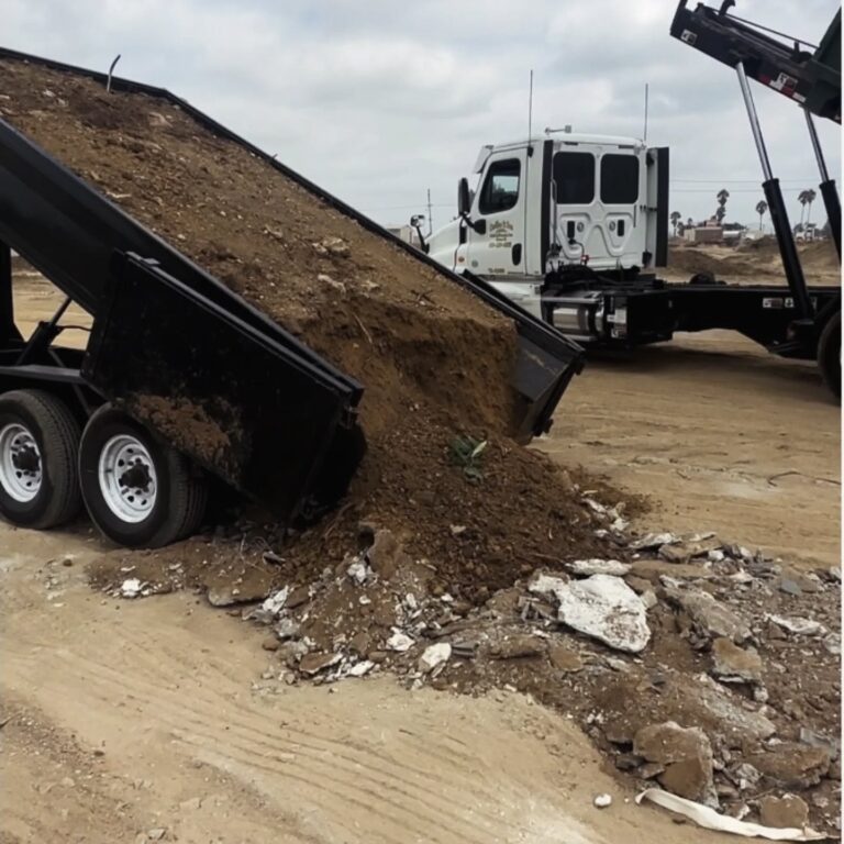 A dark gray dump truck with a white cab raises its bed, dumping a pile of brown soil and construction debris onto sandy ground. Chunks of white and gray concrete rubble mix with the spilled dirt. A flatbed truck with a raised hook lift system stands in the background under an overcast sky.