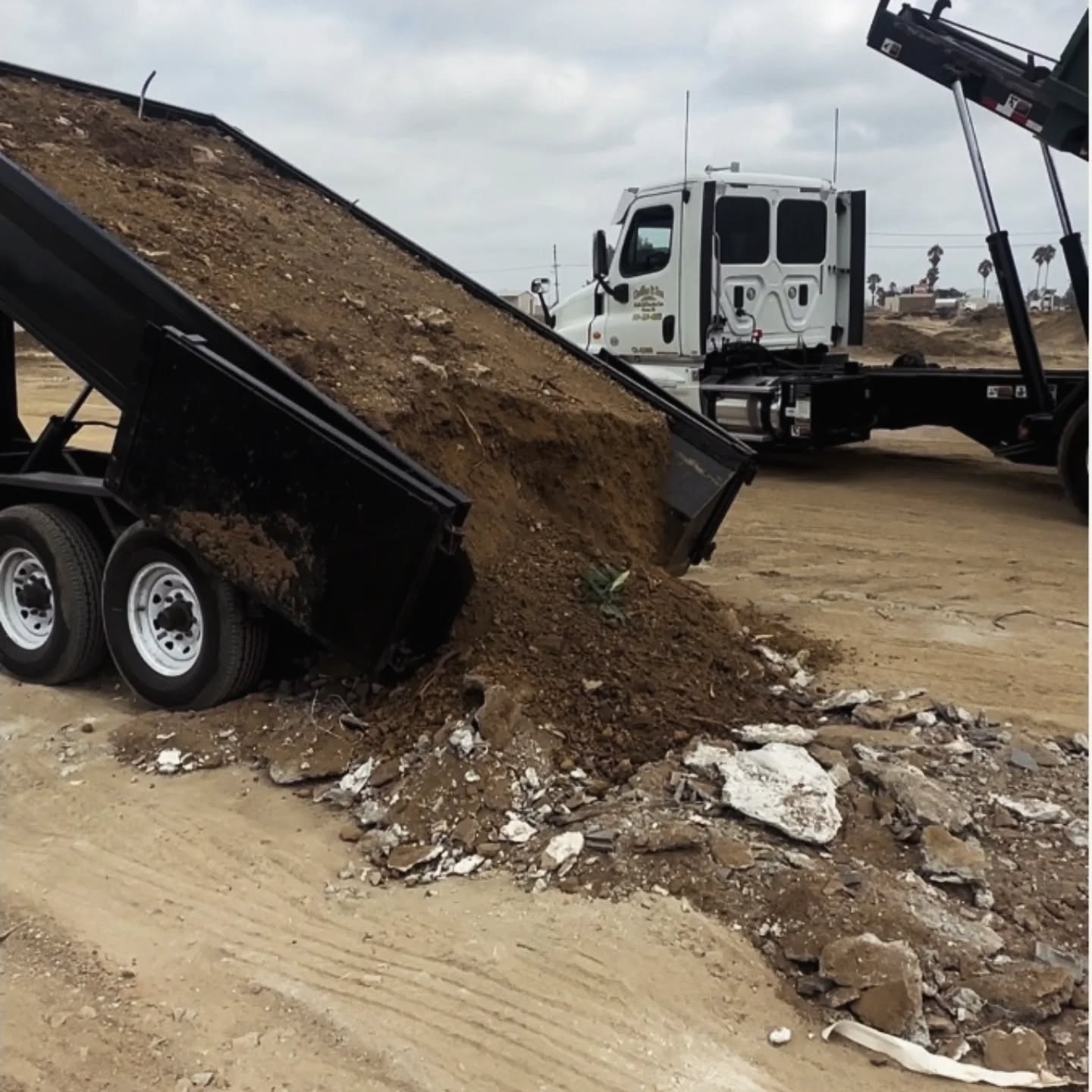 A dark gray dump truck with a white cab raises its bed, dumping a pile of brown soil and construction debris onto sandy ground. Chunks of white and gray concrete rubble mix with the spilled dirt. A flatbed truck with a raised hook lift system stands in the background under an overcast sky.