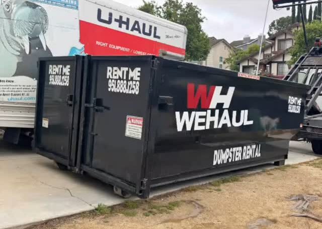 A large black dumpster with the rental company name "WH WEHAUL DUMPSTER RENTAL" printed in white and red lettering is parked on a paved driveway. The dumpster also features "RENT ME" and phone numbers on its side, along with a warning label. A white U-Haul moving truck is partially visible behind it. In the background are houses and green trees beneath an overcast sky.