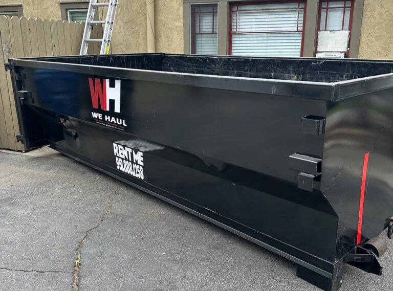 Large empty black dumpster parked on an asphalt driveway, with red and white "WH WE HAUL" logo and "RENT ME 951.888.1258" phone number on its side, light-colored building, wooden fence, and ladder in the background.