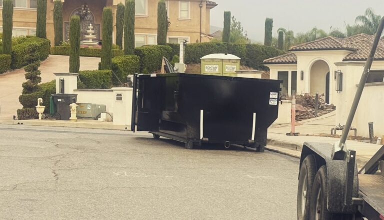 A large black dumpster on a residential street blocks the view of a house under renovation, with an ornate multi-story home, fountain, manicured landscaping, green portable toilets, and an orange construction cone visible nearby.