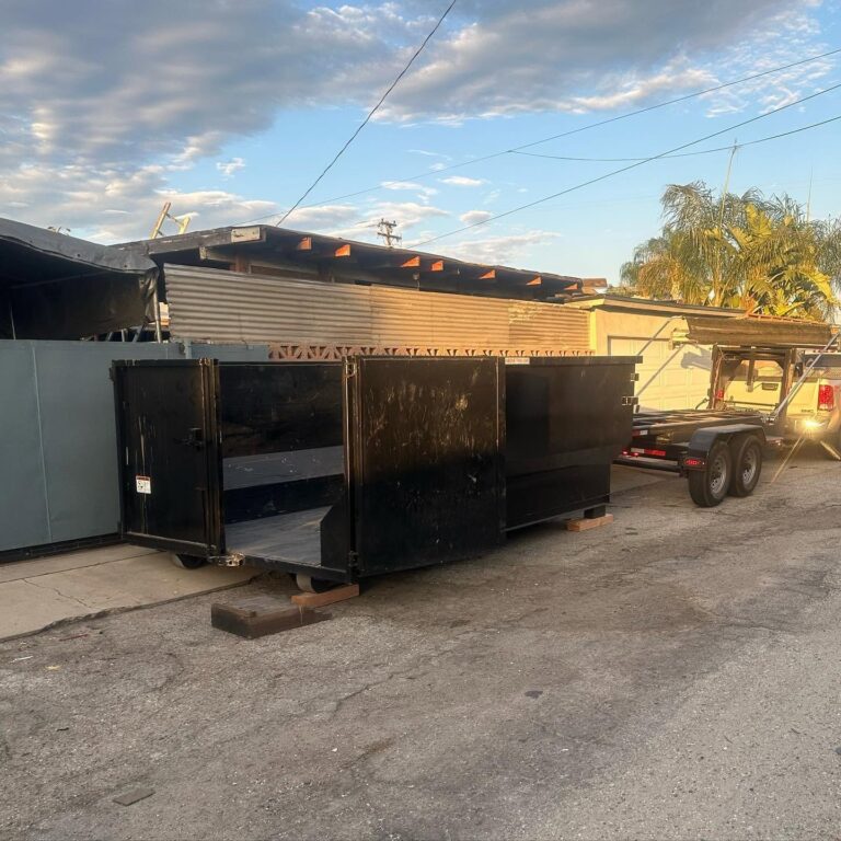 A large black roll-off dumpster with a hinged door sits on concrete, propped on wooden blocks, beside a two-axle trailer hitched to a silver pickup truck under a clear blue sky with scattered clouds, palm trees in the distance.
