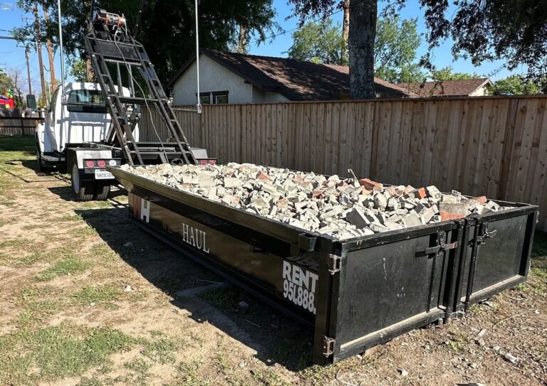 A white dump truck backs up to a black dumpster overflowing with broken concrete and red bricks on dry grass. A wooden fence and house sit in the background under a bright, clear sky.